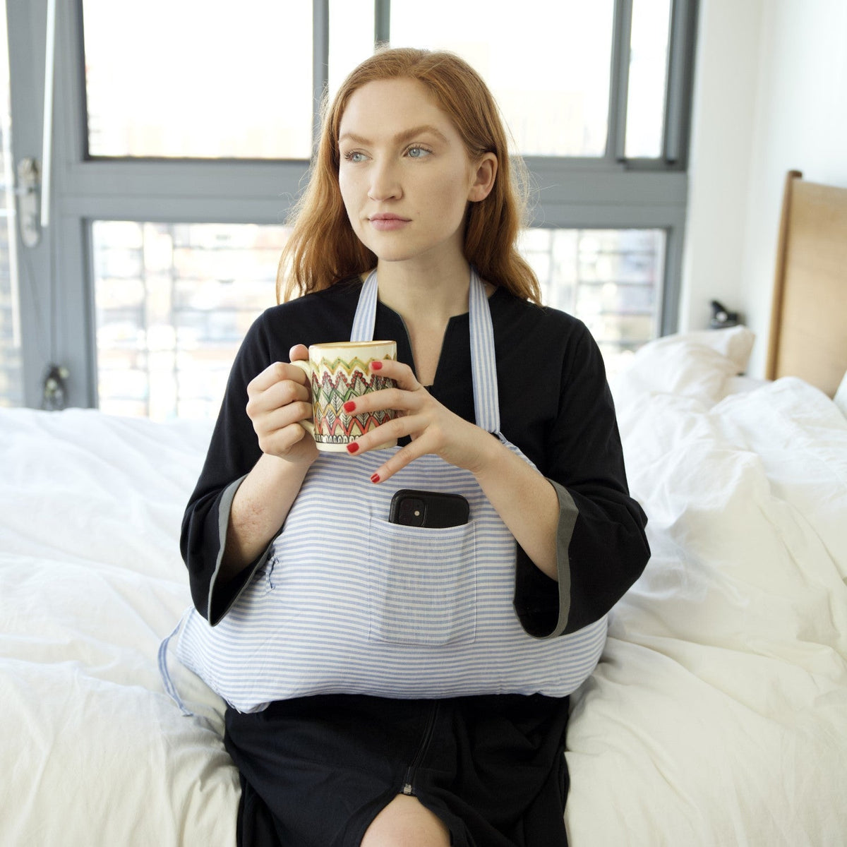 Woman wearing an Inspired Comforts post-mastectomy recovery robe with drain pockets, sitting on a bed with a mug of coffee